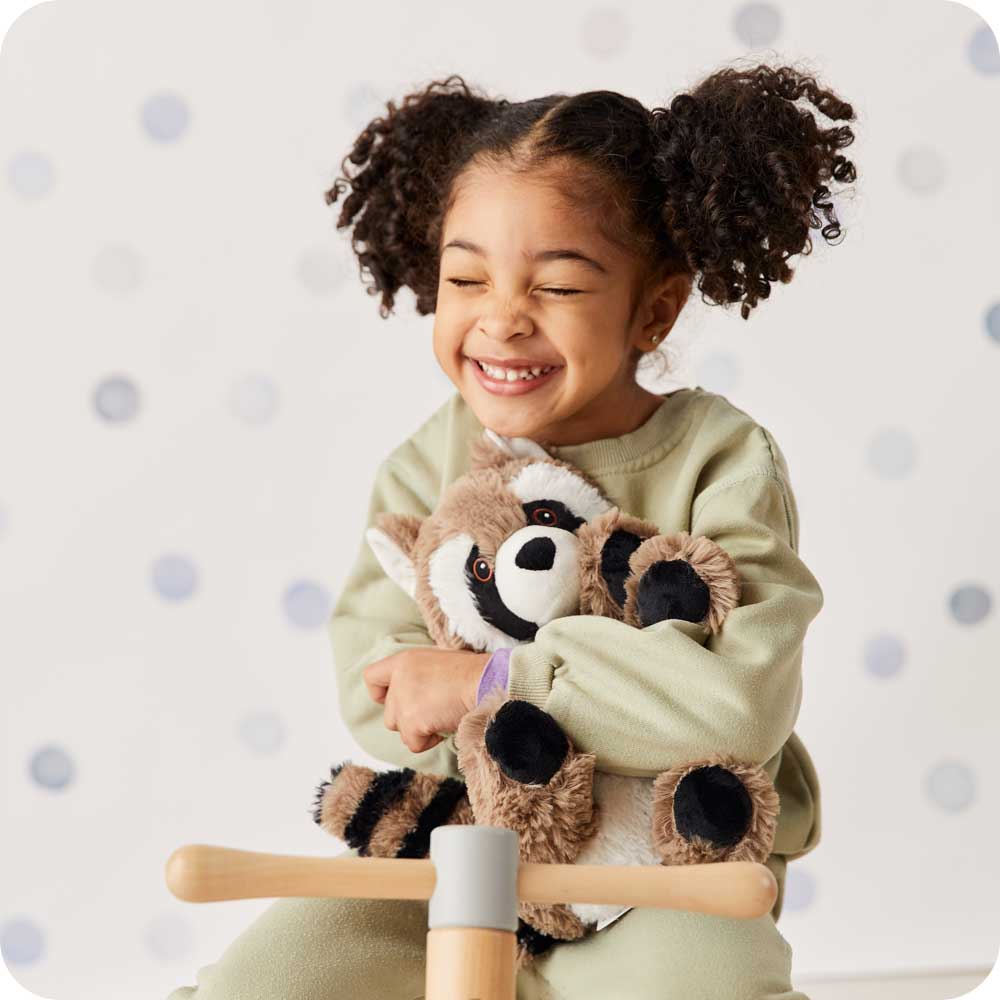 Image 104: Little girl smiling and holding Cute Weighted Lavender Scented Raccoon Stuffed Animal Heating Pad Warmies