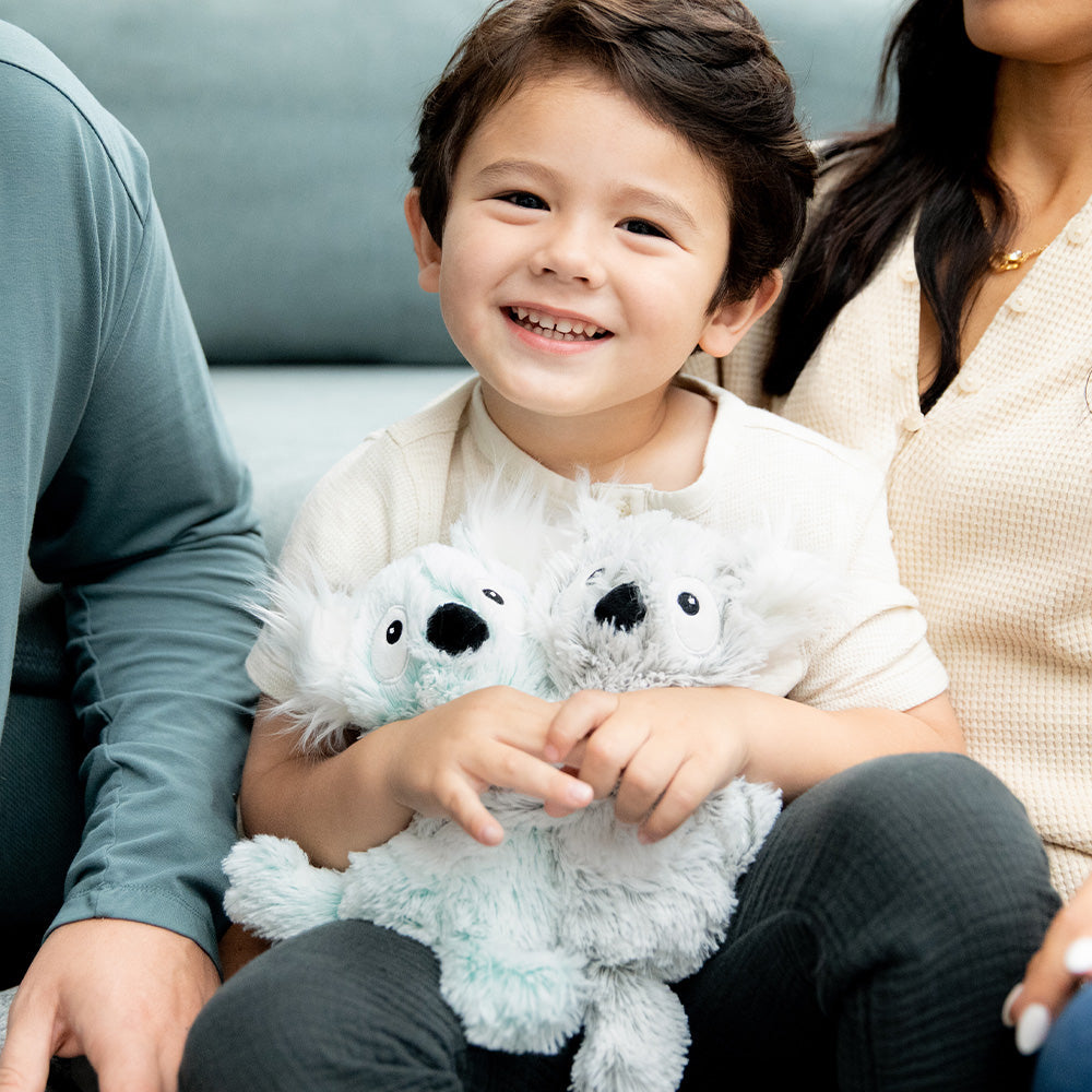 Image 101: Little boy sitting down and smiling while holding Soft Weighted Lavender Scented Koala Hugs Plush Warmies